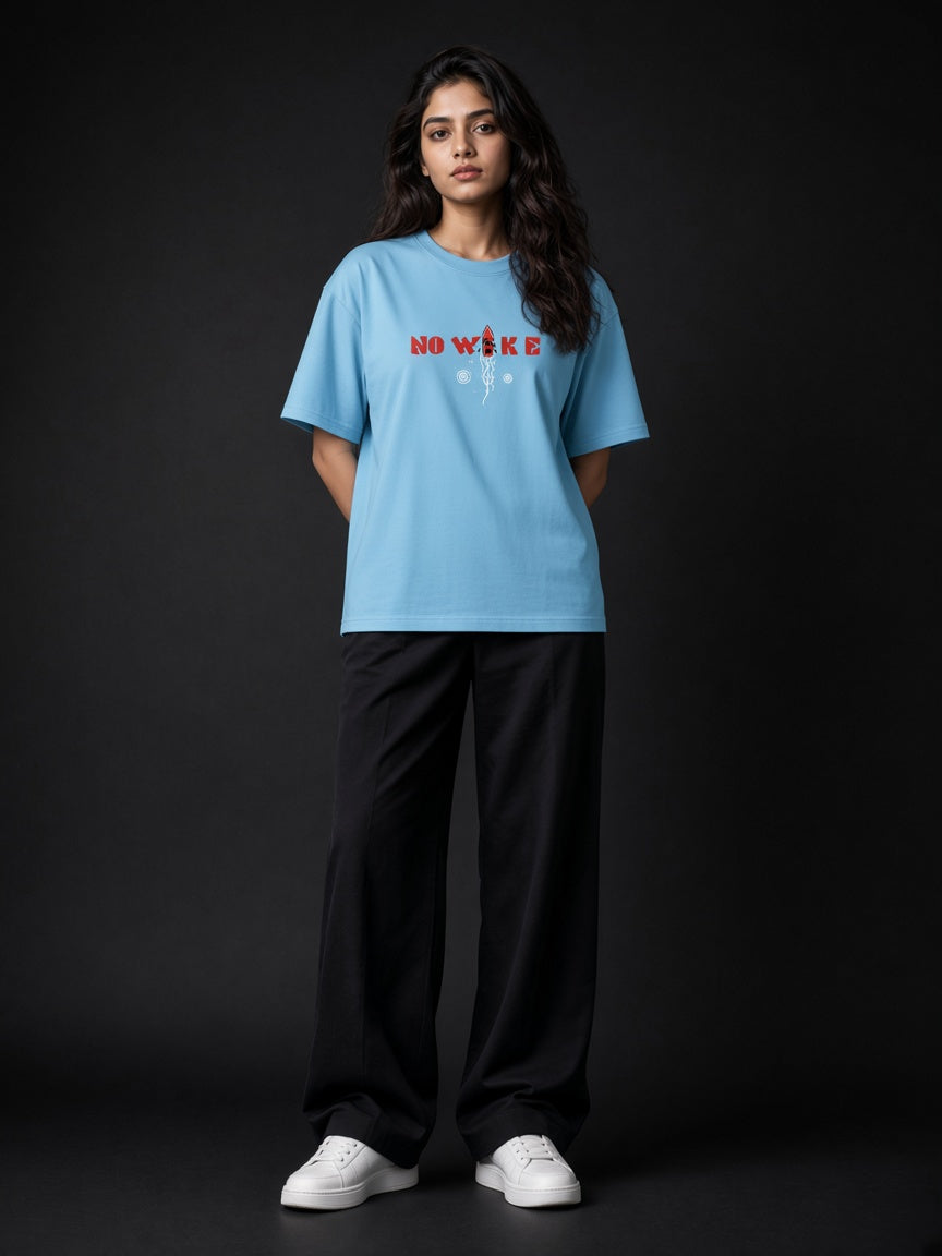 A woman modeling a light blue graphic t-shirt with 'NO WAKE' in red and white text, paired with black wide-leg pants and white sneakers, set against a dark studio background.