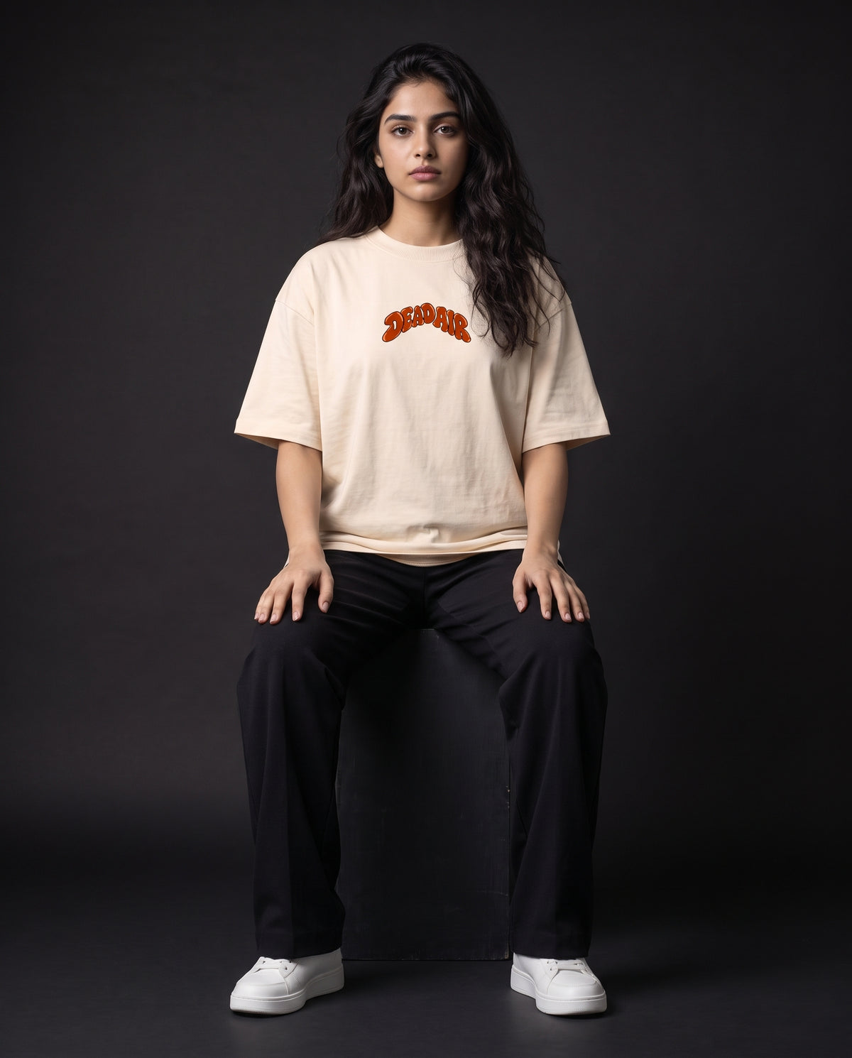 A woman sits on a dark studio block wearing an oversized cream-colored t-shirt with a red and orange 'DEADAIR' graphic on the chest, paired with black pants and white sneakers.