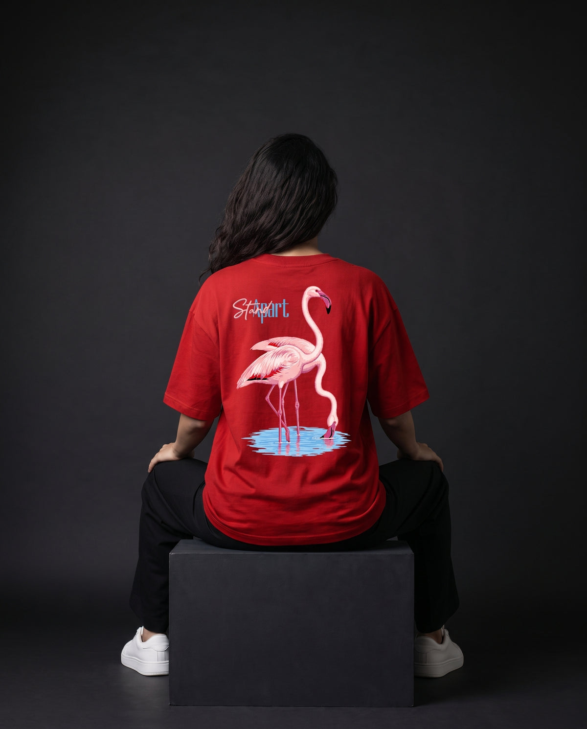 A person sitting on a black block, viewed from the back, wearing a red t-shirt with a large pink flamingo graphic and the text 'Sapient' on the back, paired with black pants and white sneakers against a dark studio background.