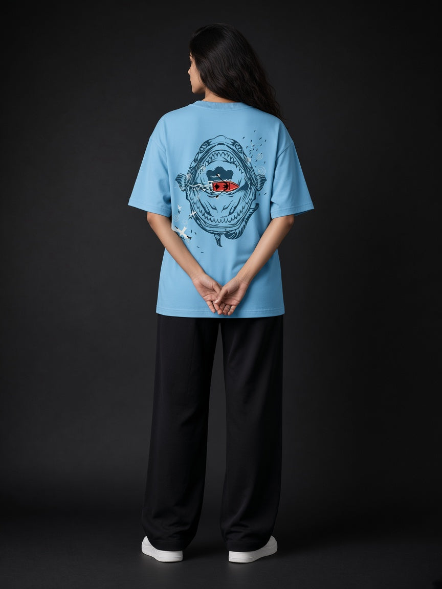A model viewed from behind wearing a light blue oversized t-shirt with a large graphic print on the back, paired with black wide-leg pants and white sneakers, set against a dark studio background.