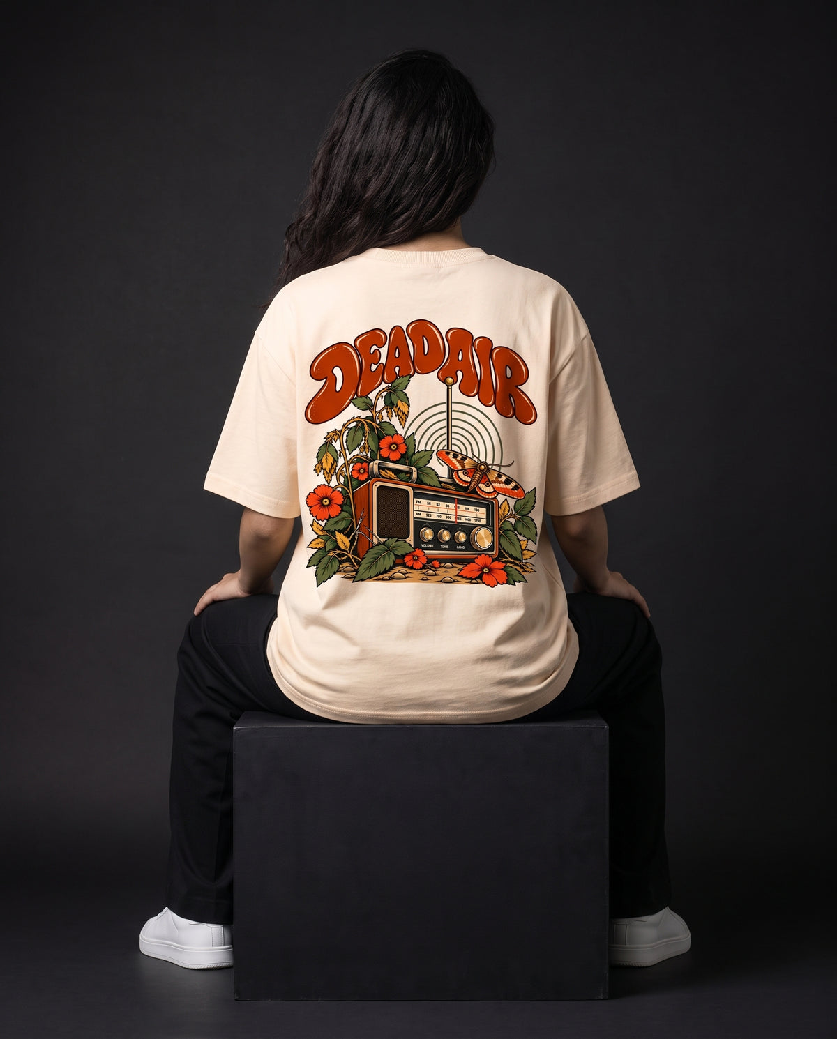 A person sitting on a black block, viewed from the back, wearing a cream-colored t-shirt with a large retro graphic on the back. The graphic features the word 'DEADAIR' in orange-red bubble letters above an illustration of a vintage radio surrounded by red flowers and green leaves.