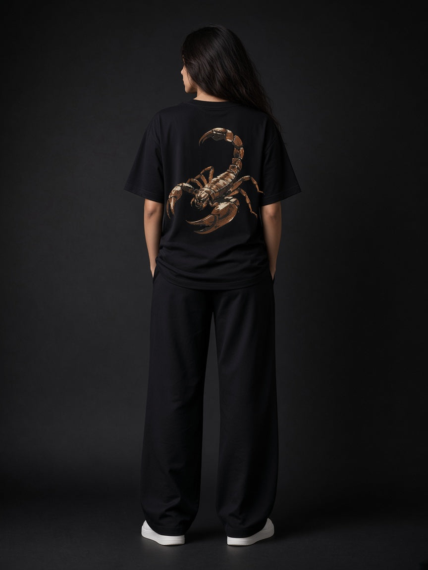 A model viewed from behind wearing a black t-shirt with a large brown scorpion graphic on the back, paired with black wide-leg pants and white sneakers, set against a dark studio background.