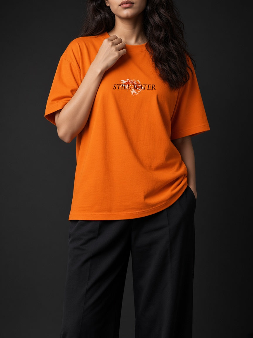 A woman wearing a bright orange short-sleeve t-shirt with a black 'STILL WATER' graphic and a white dragon illustration on the chest, paired with black pants, set against a dark studio background.