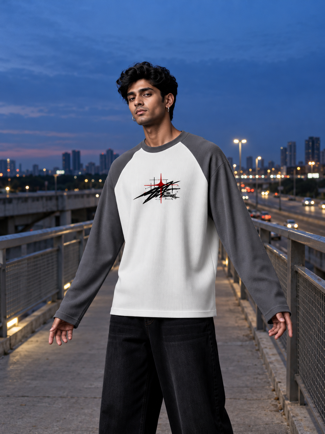A man wearing a raglan long-sleeve shirt with a white body, gray sleeves, and a black and red graphic on the chest, standing on an outdoor walkway at dusk.