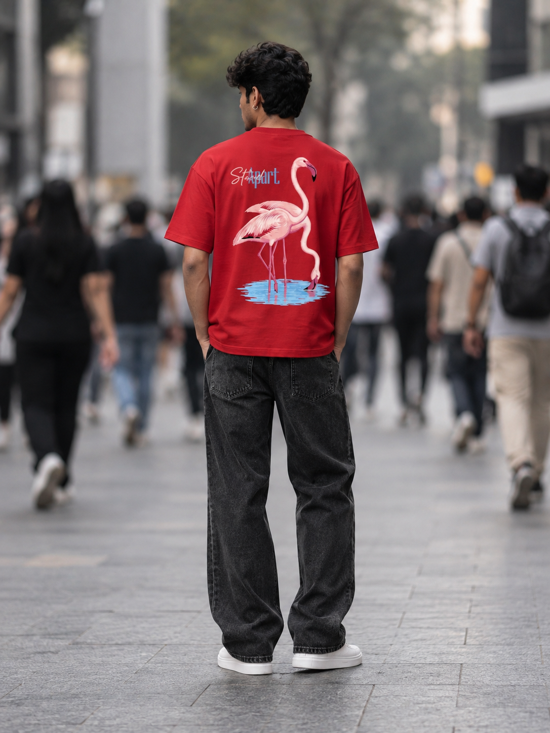 A man viewed from behind wearing a bright red t-shirt with a large pink flamingo graphic and the text 'Shapell' on the back, paired with dark gray jeans and white sneakers, standing in a busy outdoor city walkway.
