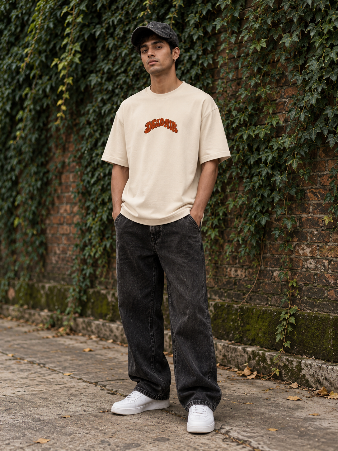 A man wearing a cream-colored t-shirt with a red graphic, dark gray baggy jeans, white sneakers, and a black cap, standing outdoors against a brick wall covered in green ivy.