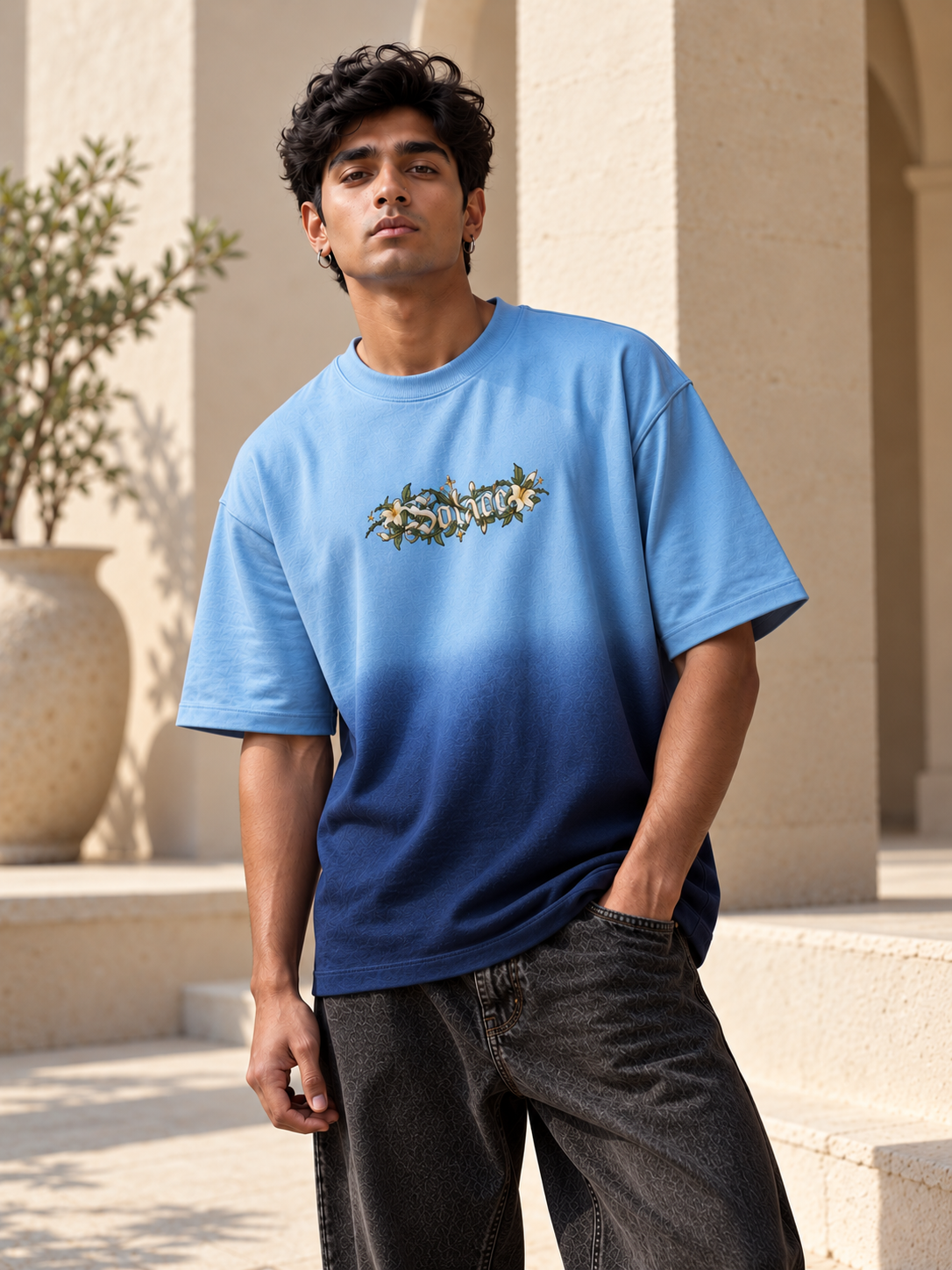 A man wearing a light blue to dark blue gradient t-shirt with a floral graphic on the chest, paired with dark gray jeans, standing outdoors in front of beige stone columns.