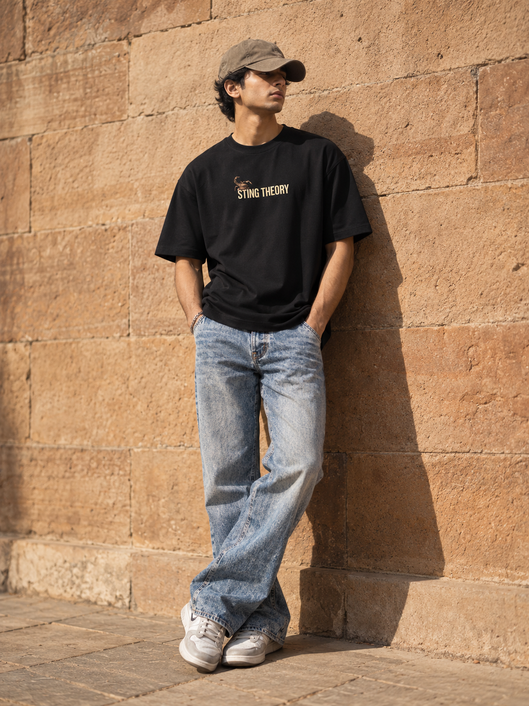 A man wearing a black graphic t-shirt with 'STING THEORY' printed in gold on the chest, paired with light-wash blue jeans, white sneakers, and a beige baseball cap, leaning against a textured tan stone wall.