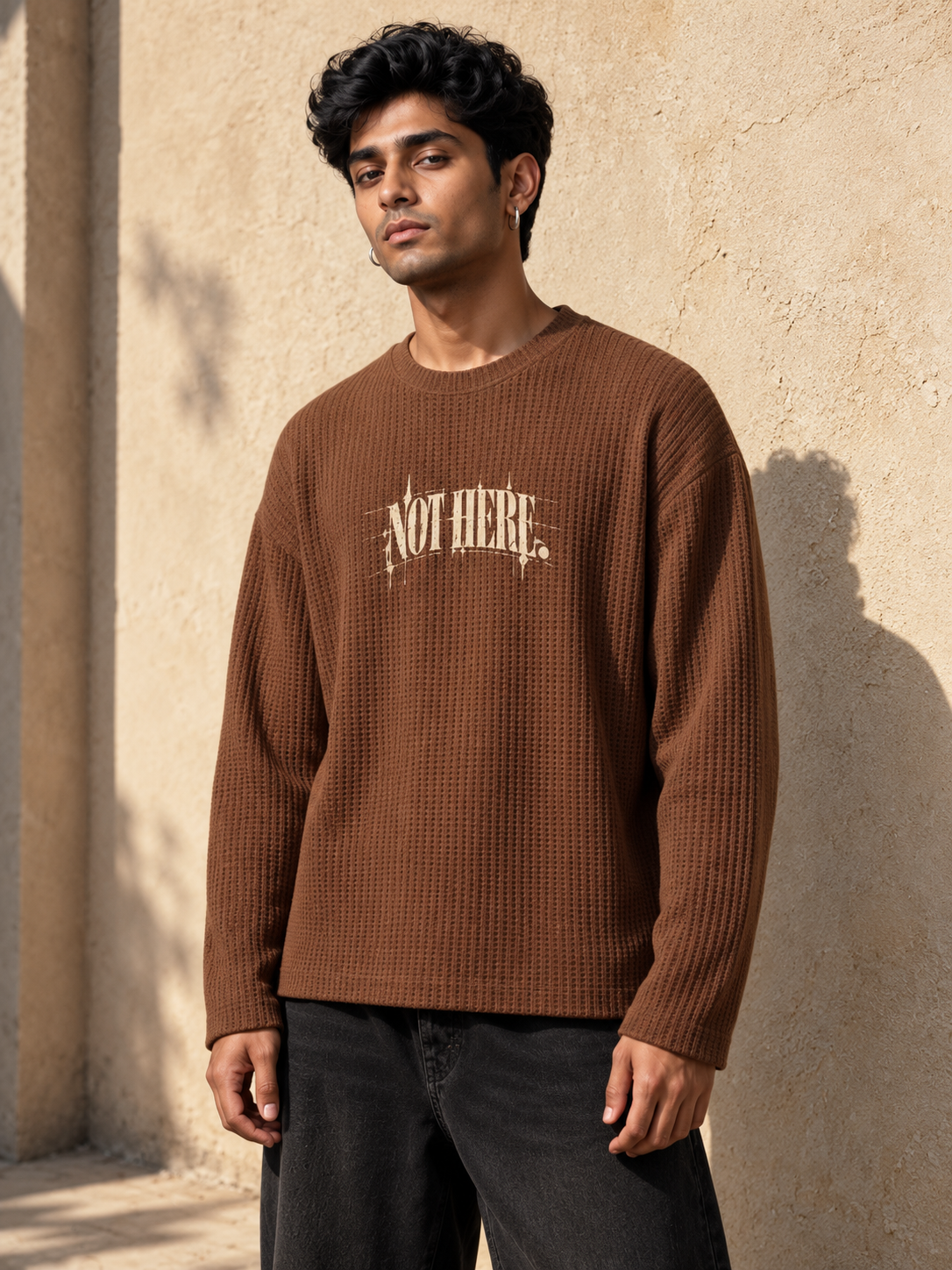 A man wearing a brown waffle-knit long-sleeve sweater with 'NOT HERE' printed in cream across the chest, standing against a textured beige wall.
