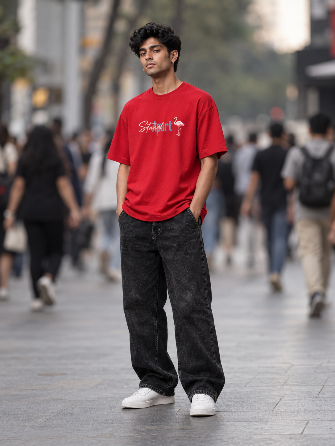 A man standing on a city street wearing a bright red graphic t-shirt with a white and blue script logo and a pink flamingo, paired with dark gray baggy jeans and white sneakers.